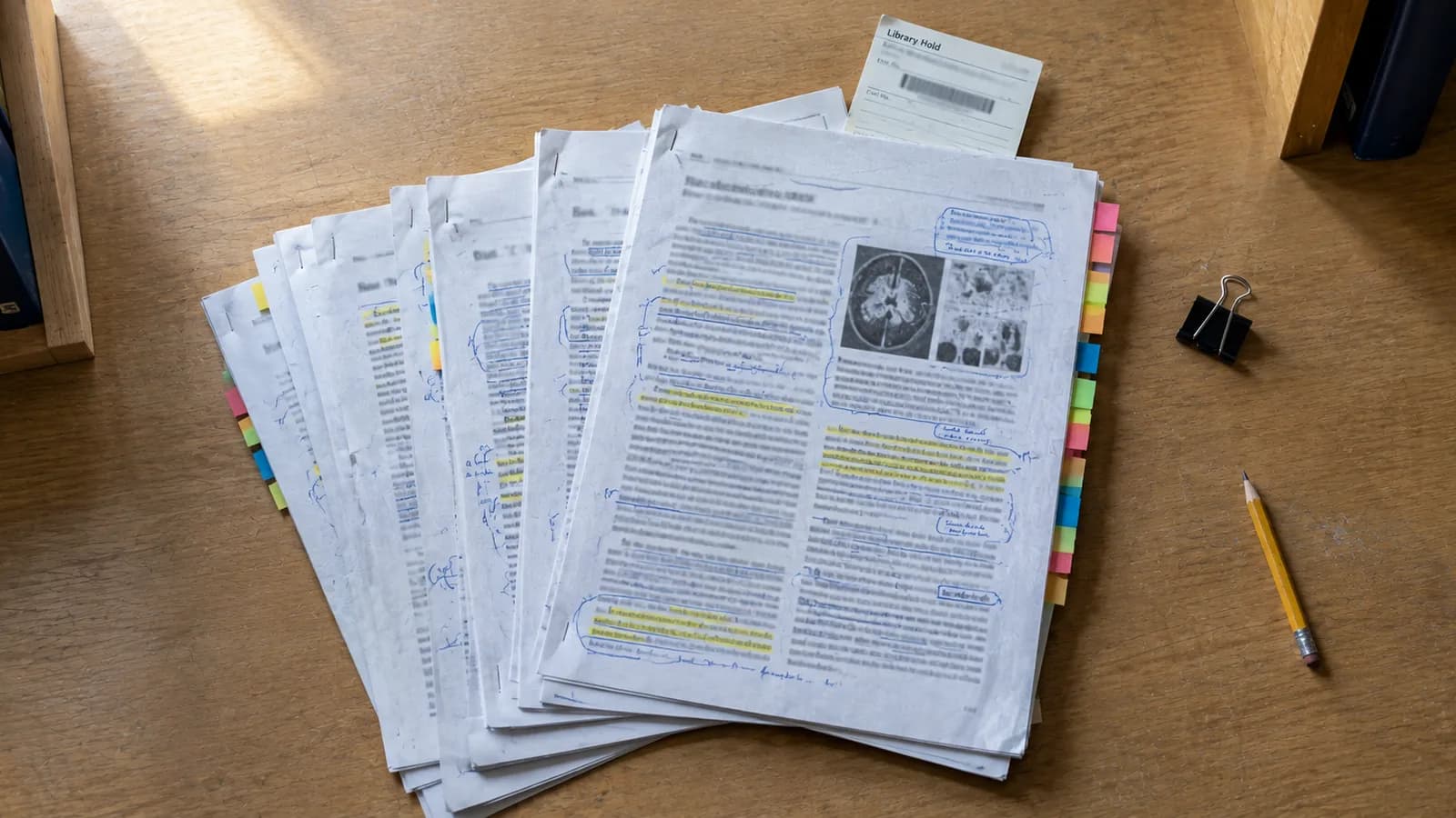 A fanned stack of stapled, photocopied journal articles on a wooden library desk, annotated throughout in blue pen and yellow highlighter, with colour-coded sticky tabs along one edge, a Library Hold slip and barcode resting on top, and a binder clip and a short pencil beside them.