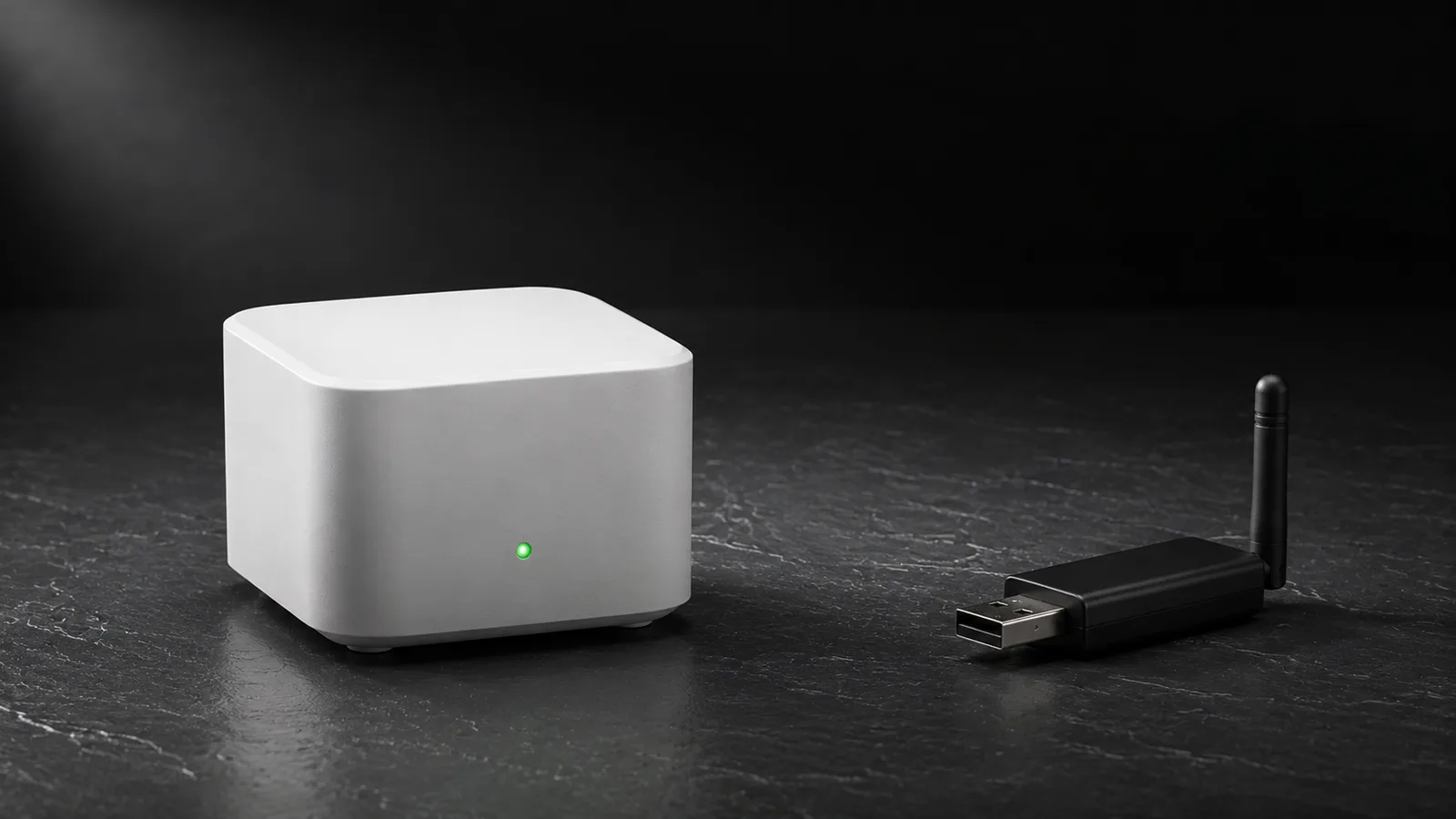 A small white cube smart home hub with a single green status light on a dark slate surface, and a separate black USB dongle with a stub antenna lying a few inches to its right, a clear gap of empty surface between them.