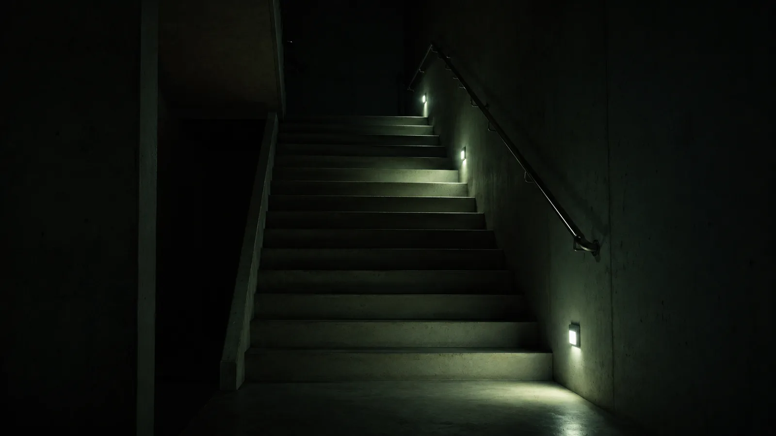 A dark concrete stairwell lit only by three small wall-mounted emergency lamps, their pale pools of light picking out the steps and handrail while the rest of the space stays in shadow.